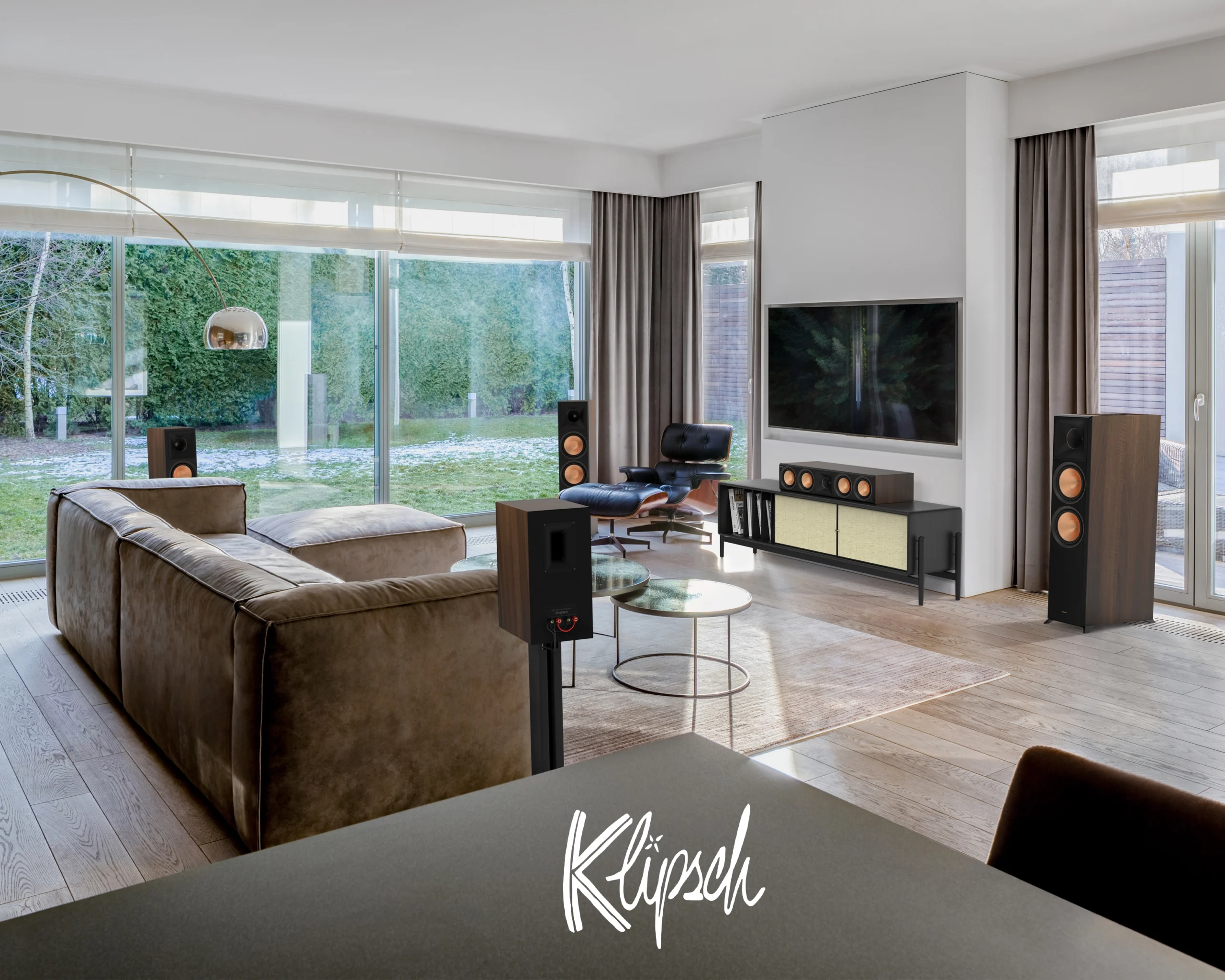 A Klipsch system in a garden-facing living room: floorstanding speakers either side of a wall-mounted television, with a centre speaker on a low cabinet.