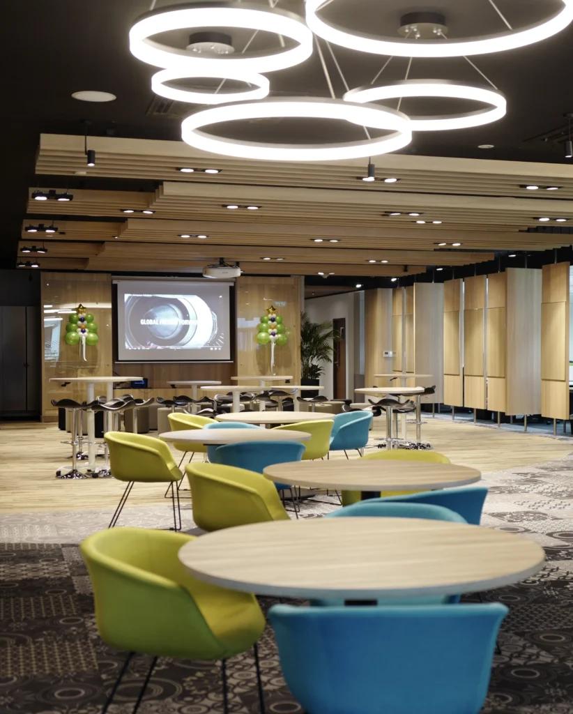 A corporate event space with ring pendant lights, a projection screen, high tables and green and blue tub chairs.