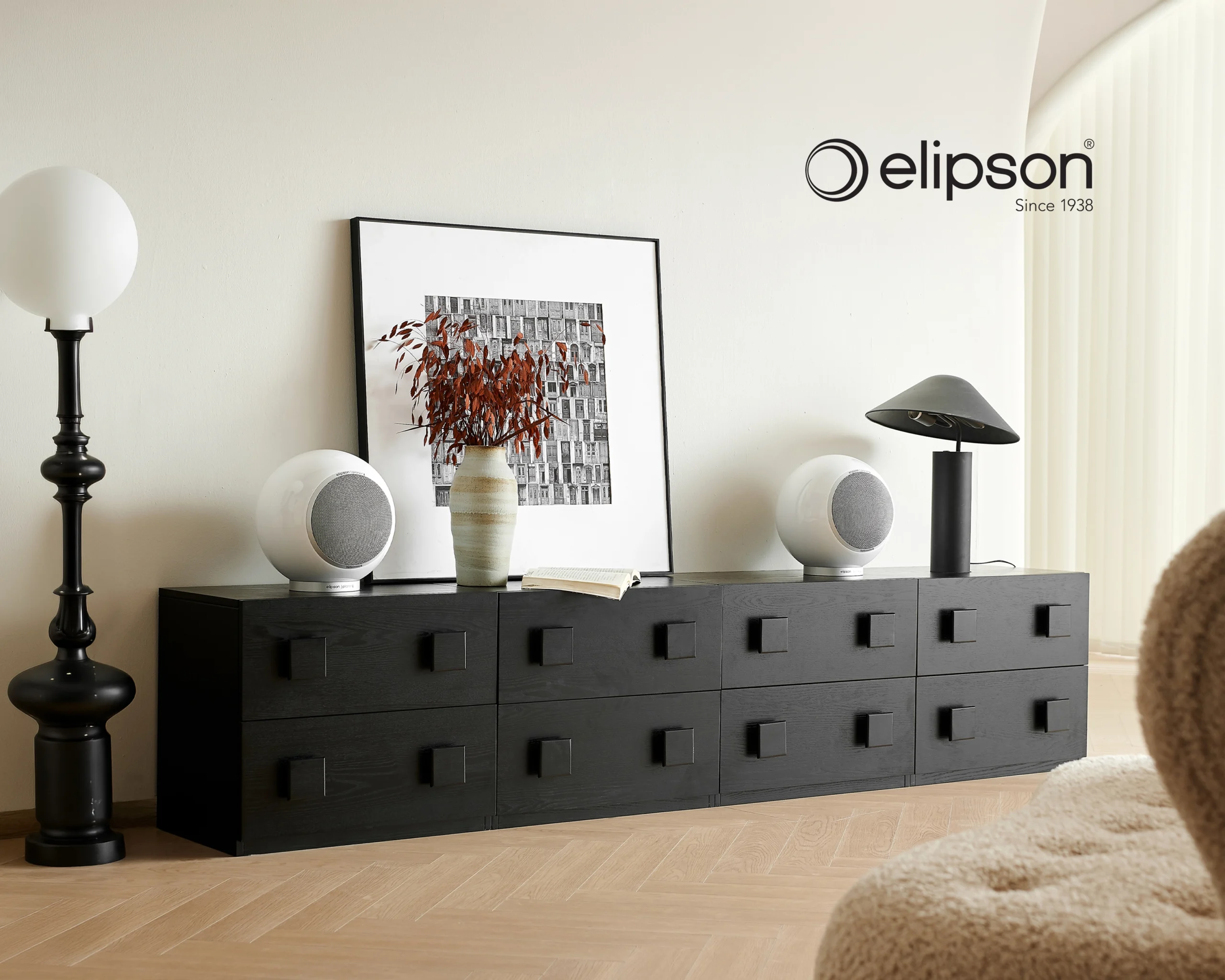 Two white spherical Elipson loudspeakers on a black sideboard, beside a framed print and a table lamp.