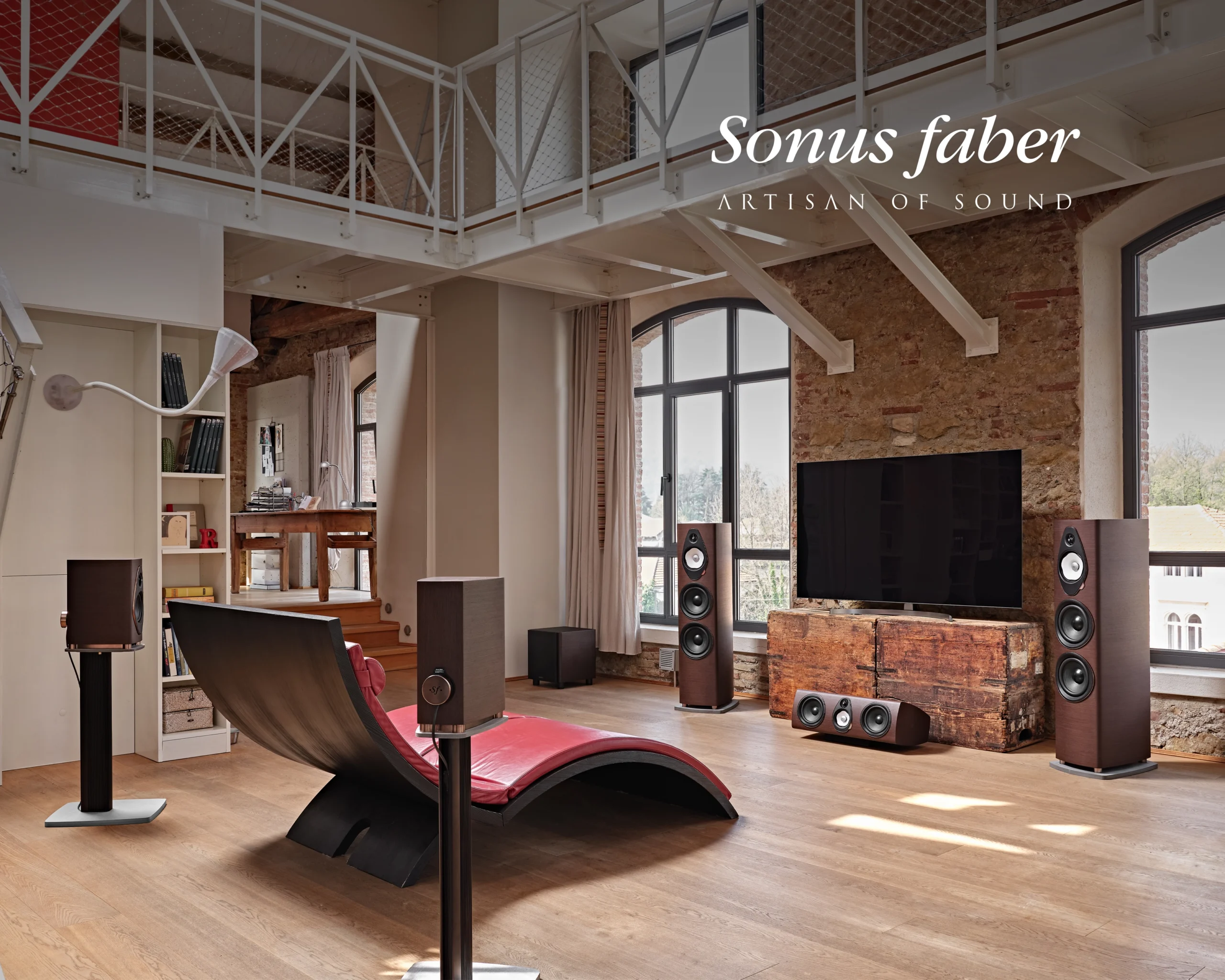A Sonus Faber system in a converted loft with exposed brick and a mezzanine, arranged around a television and a red chaise longue.