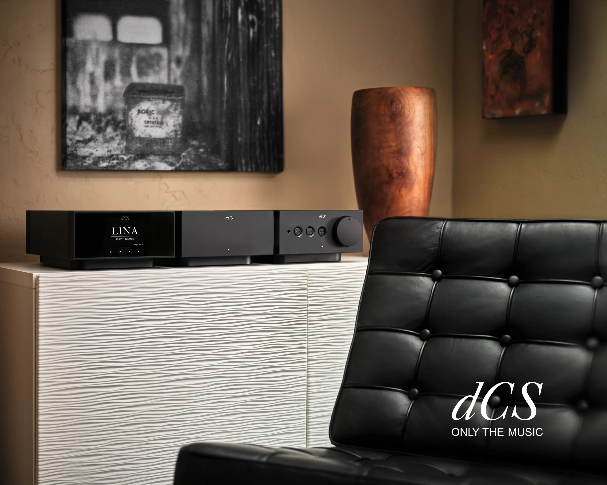 Three black dCS components on a white ribbed cabinet, the top unit's display reading LINA, beside a black leather chair.