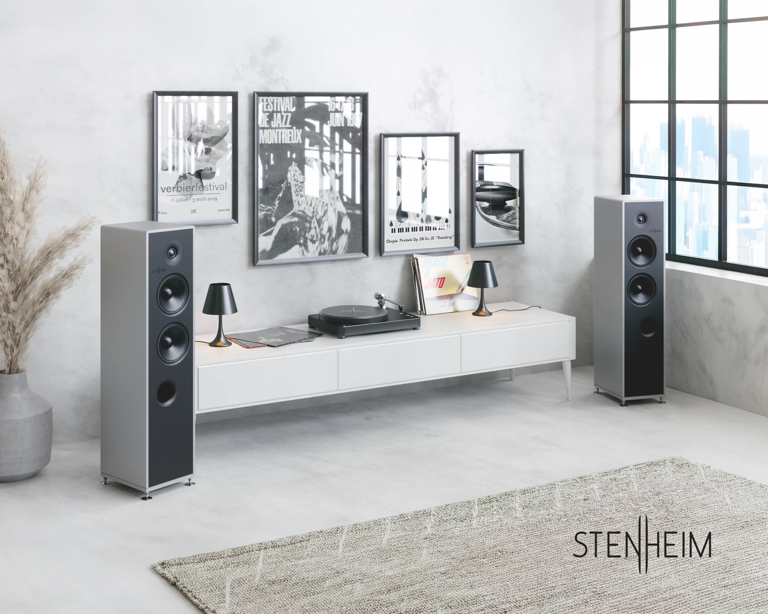 Two Stenheim floorstanding loudspeakers either side of a long white sideboard holding a turntable, below framed jazz festival posters.