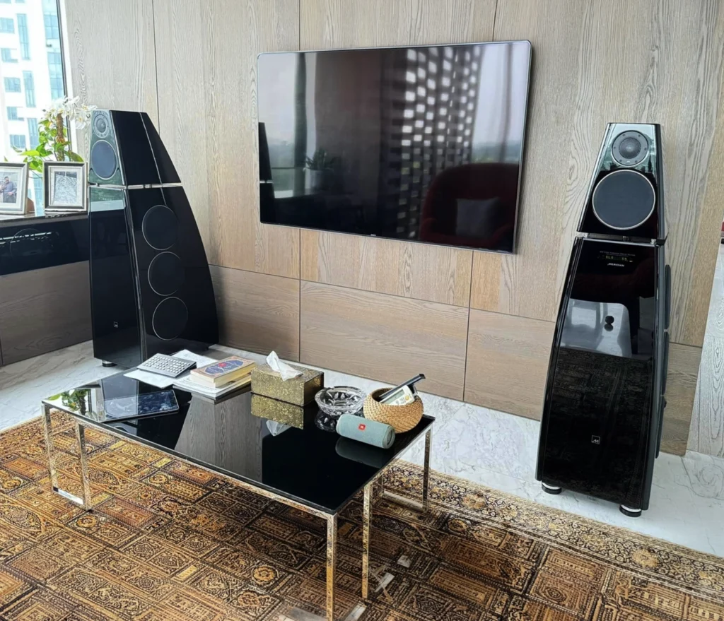 Two gloss black Meridian DSP8000 floorstanding loudspeakers either side of a wall-mounted television on an oak-panelled wall, with no cabling visible.