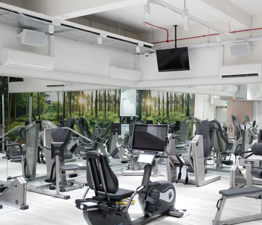 White surface-mounted loudspeakers high on the walls of an ActiveSG gym, above rows of resistance machines and exercise bikes.