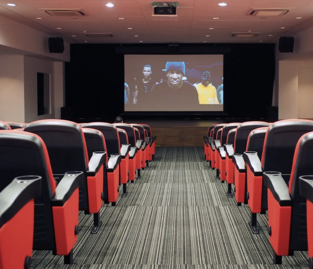 A corporate auditorium with tiered red seating facing a projection screen, and black surround loudspeakers bracketed high on the side walls.
