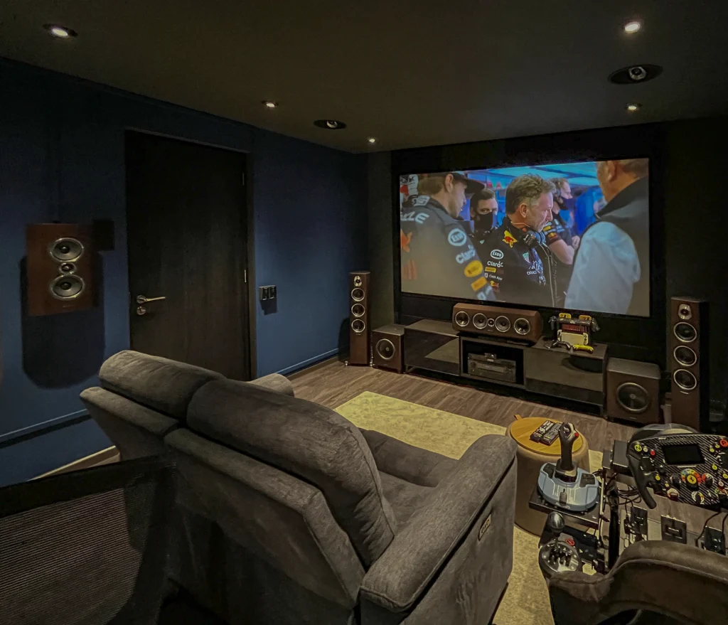 A Sonus Faber 7.2.4 private cinema in a dark blue room, with a projection screen, centre speaker and floorstanders.