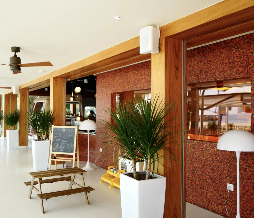 A weather-proof loudspeaker mounted on a timber column at Trapizza, a beachfront restaurant, above planters and outdoor seating.