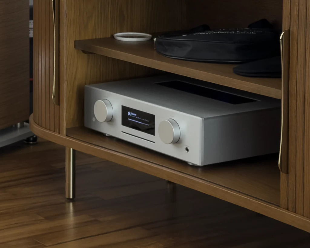 A close view of the silver AVM Evolution CS 3.3 streaming amplifier on a shelf inside the fluted walnut sideboard, its display lit.