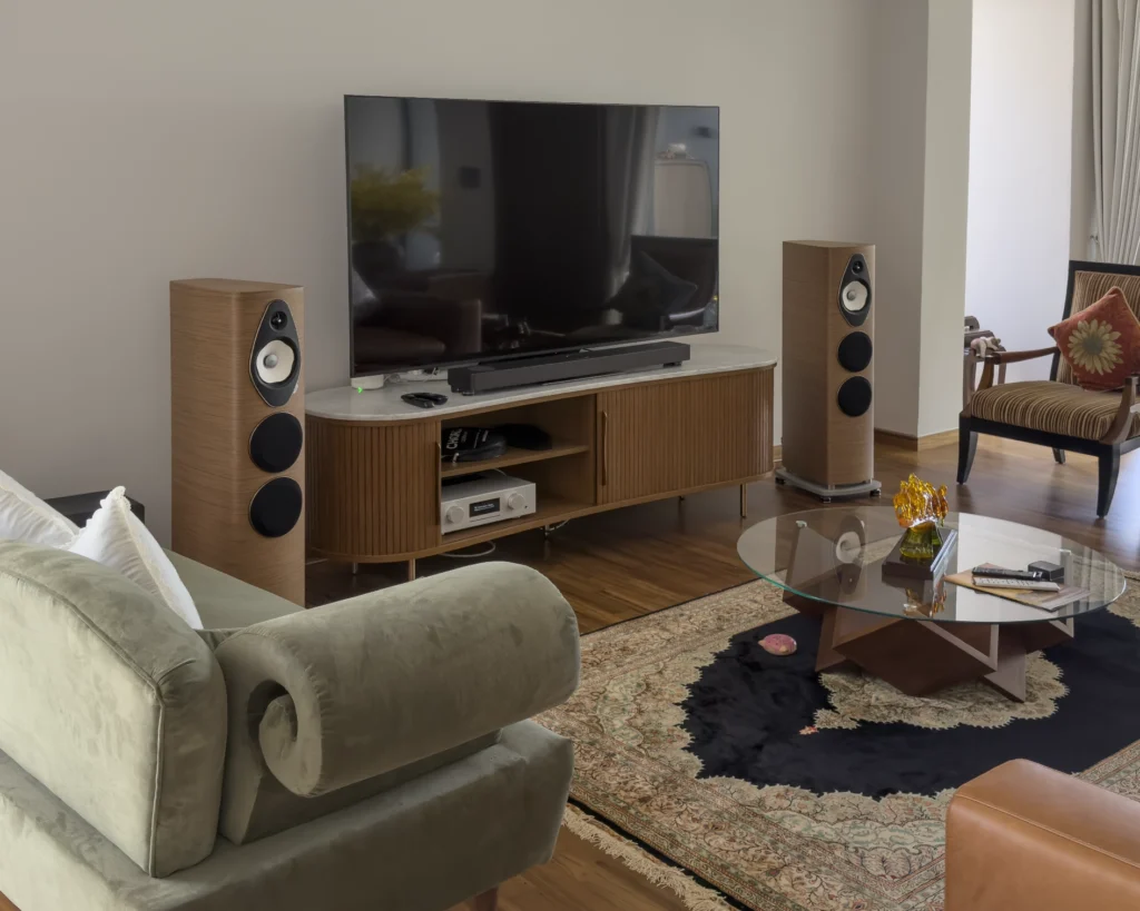 A wider view of the living room: the walnut Sonus Faber loudspeakers either side of a fluted sideboard and wall-mounted television, with a green velvet sofa and patterned rug in the foreground.
