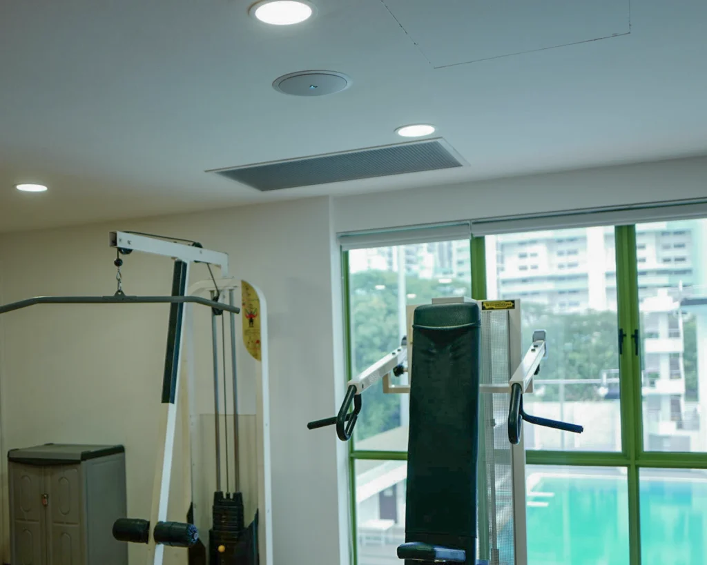Ceiling speaker and recessed lights above weight machines, with a swimming pool visible through the window