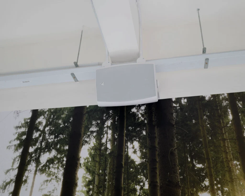 White wall-mounted speaker fixed to a ceiling beam above a forest-print wall