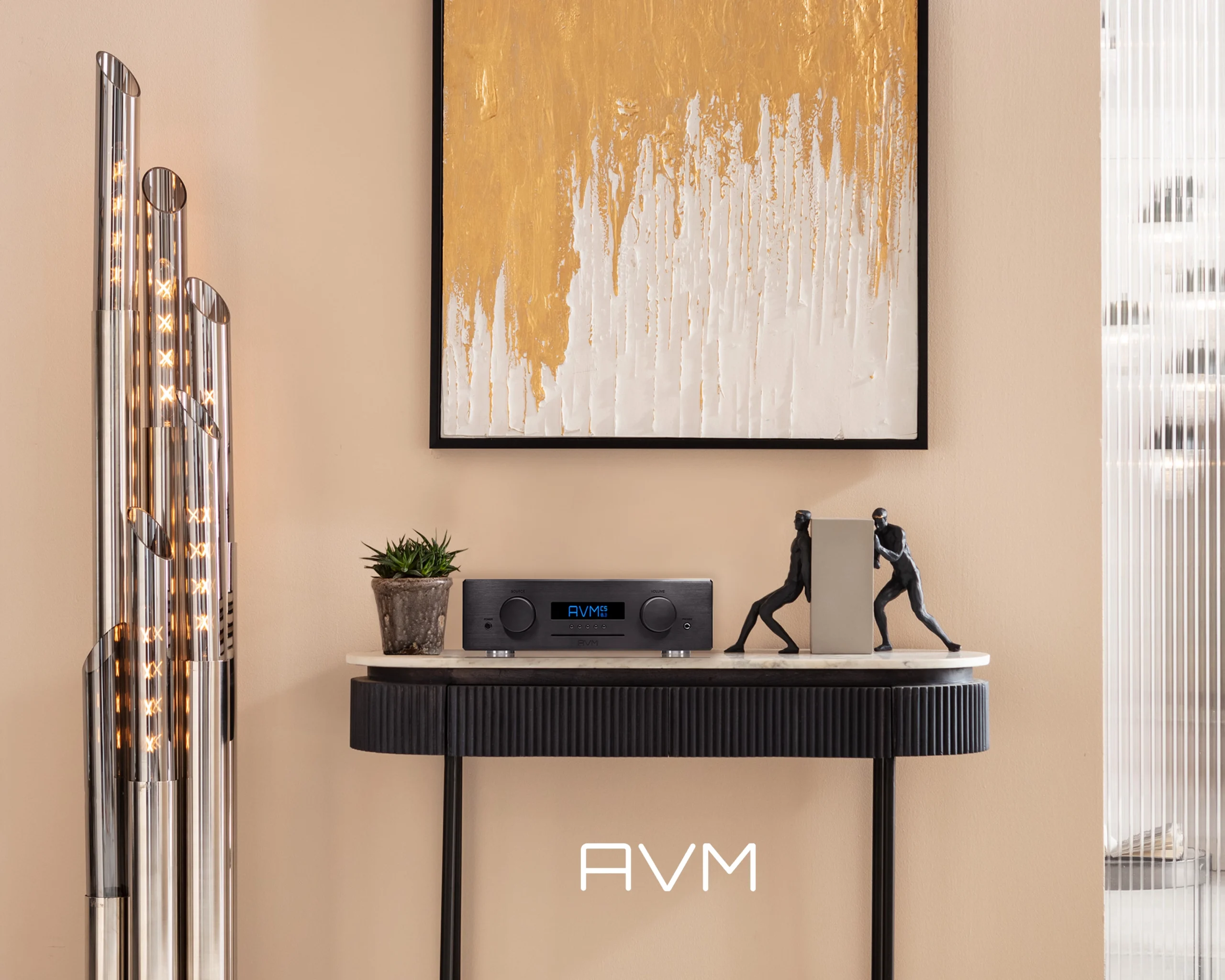 AVM integrated amplifier on a black fluted console beneath abstract gold artwork, beside a floor lamp and a sculpture.