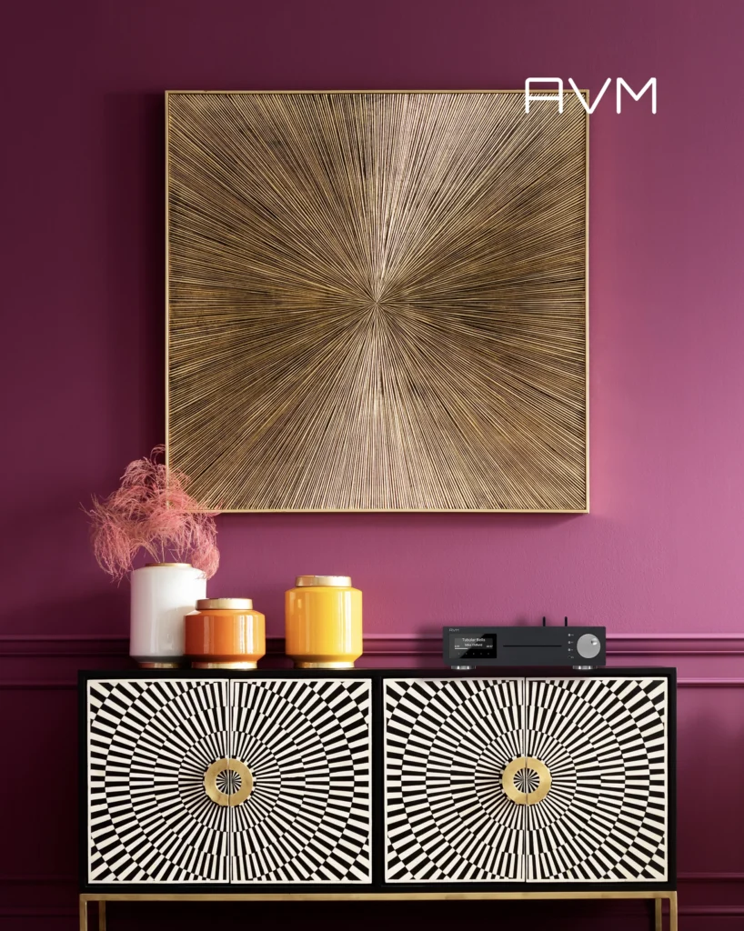 AVM streaming amplifier on a black and white patterned sideboard against a plum wall with a gold sunburst panel.