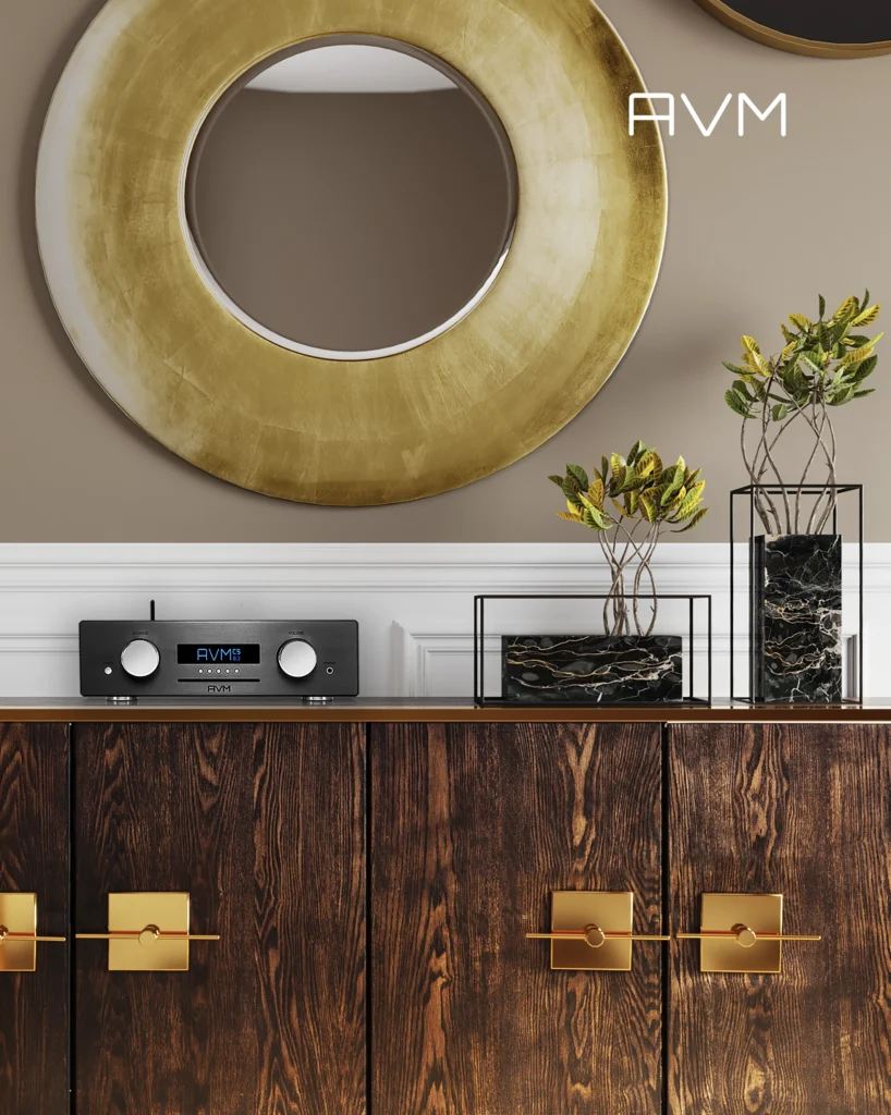 AVM integrated amplifier with a display on a dark wooden sideboard under a round gold mirror, beside a marble vase.