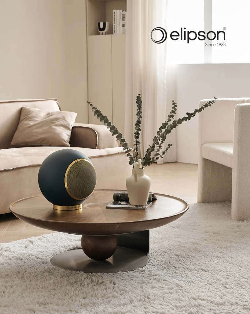 Dark blue Elipson spherical speaker with a gold base ring on a round wooden coffee table in a neutral lounge.