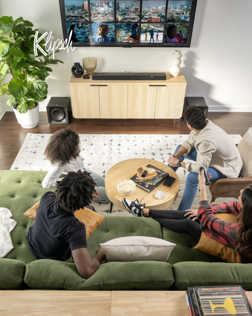 Family on a green sofa watching television, with a Klipsch soundbar and subwoofer on a light wood cabinet.