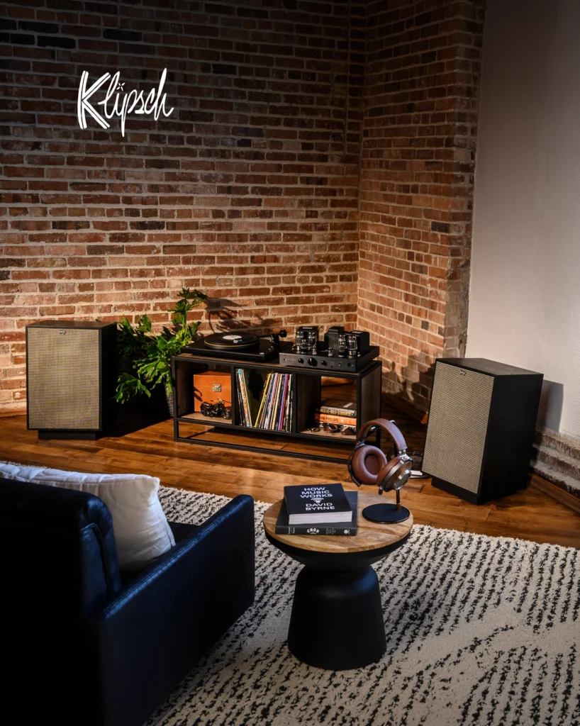 Klipsch speakers either side of a turntable and valve amplifier on a record shelf in a brick-walled room.