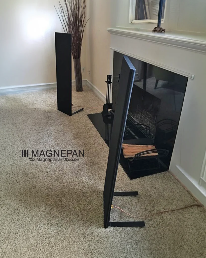 Two black Magnepan panel speakers on stands in a carpeted room with a fireplace.