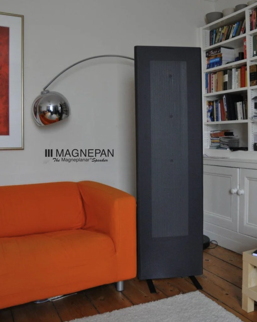 Tall black Magnepan panel speaker beside an orange sofa and a bookcase in a living room.