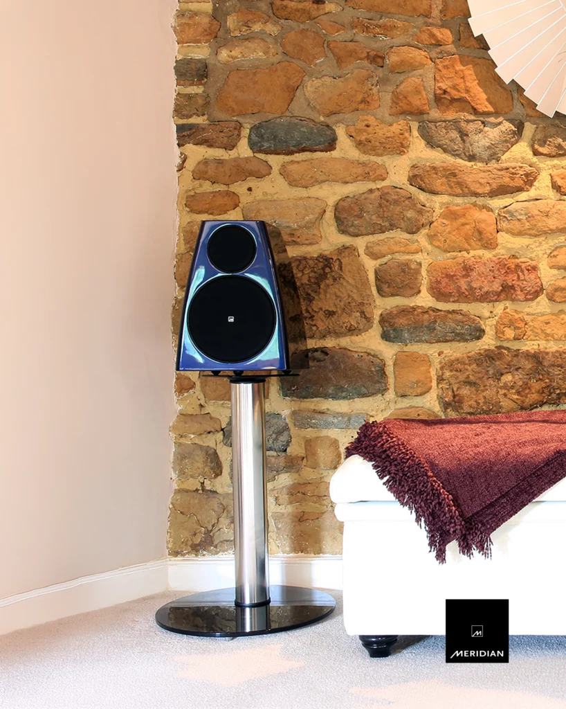 Meridian speaker in a dark blue gloss finish on a chrome stand against a stone wall, beside a white sofa.