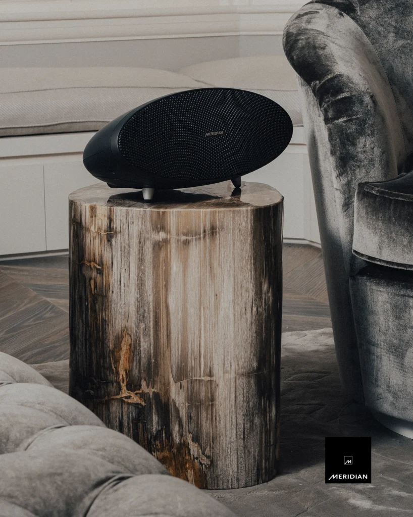 Black oval Meridian speaker resting on a petrified-wood side table between grey velvet chairs.