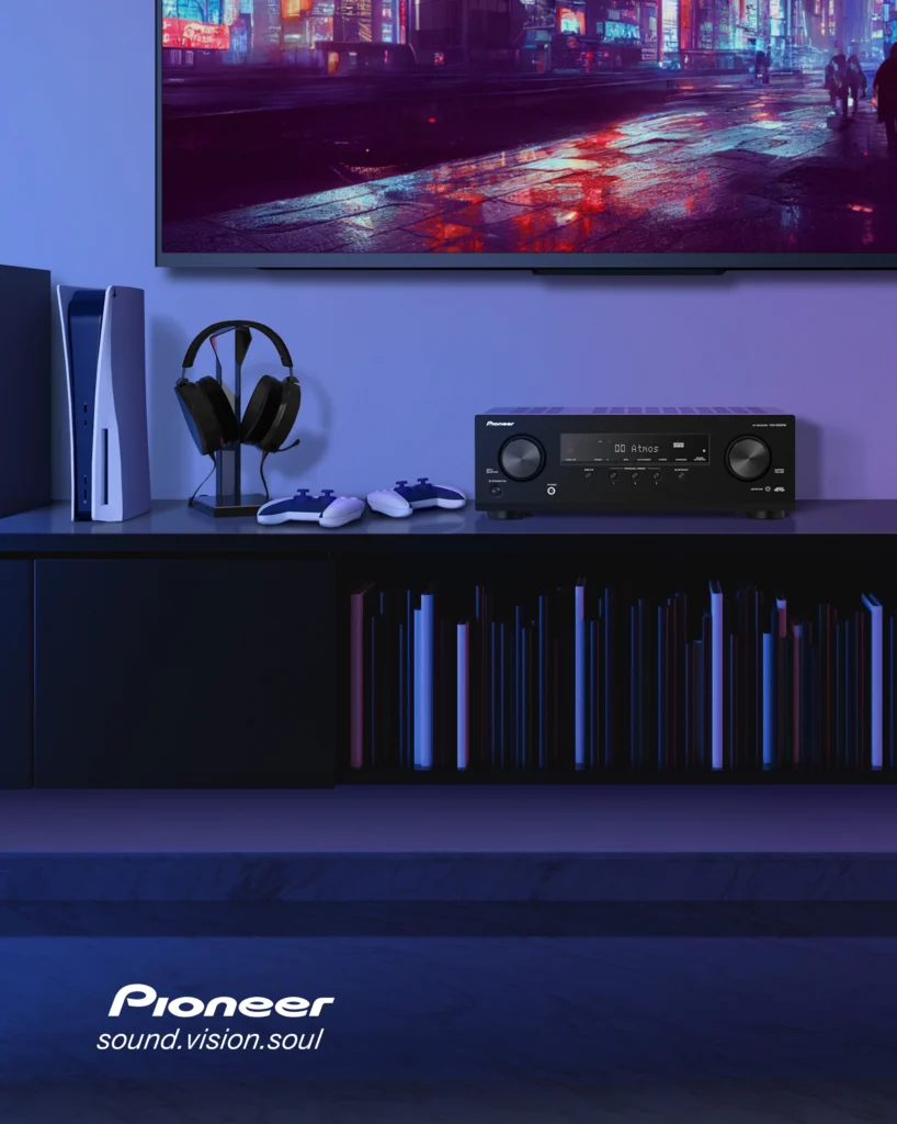 Pioneer AV receiver on a dark shelf beside a games console, headphones and controllers, beneath a television.