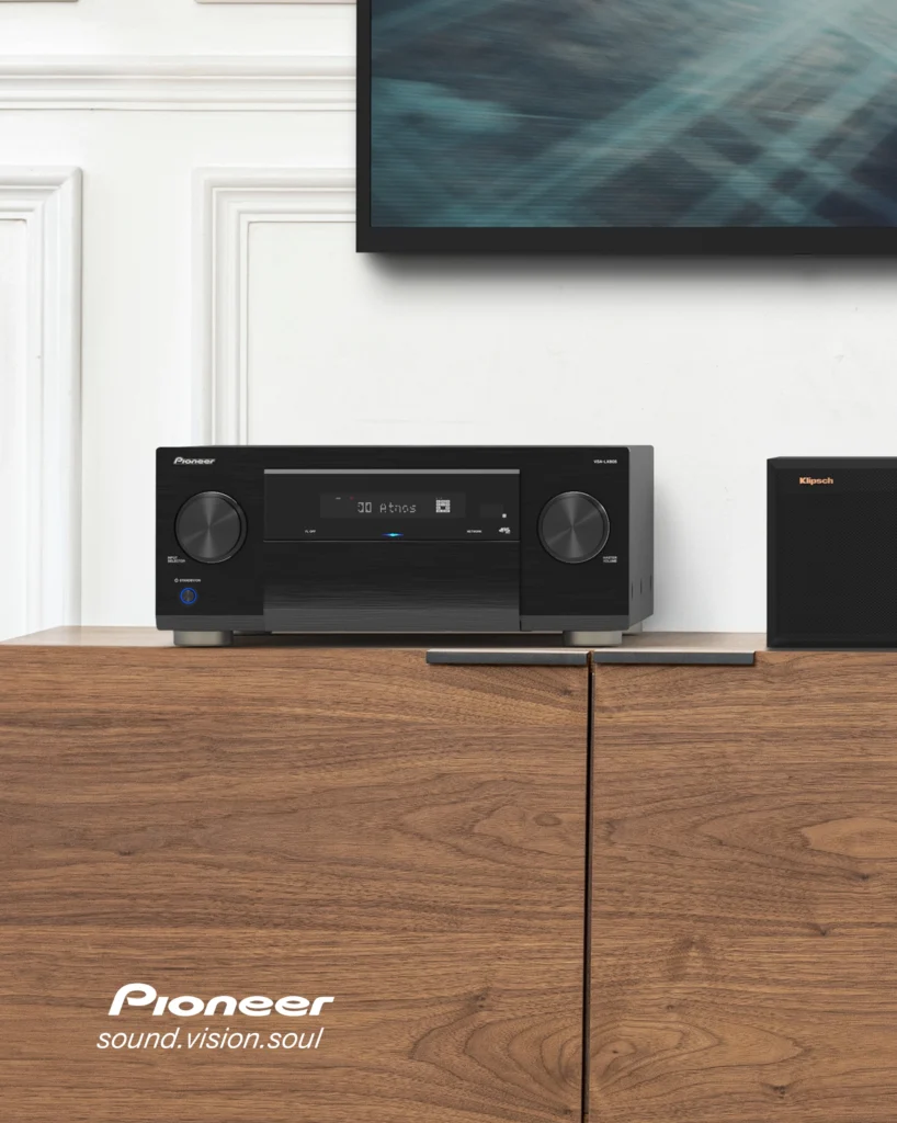 Black Pioneer AV receiver on a walnut cabinet beneath a wall-mounted television, with a speaker beside it.