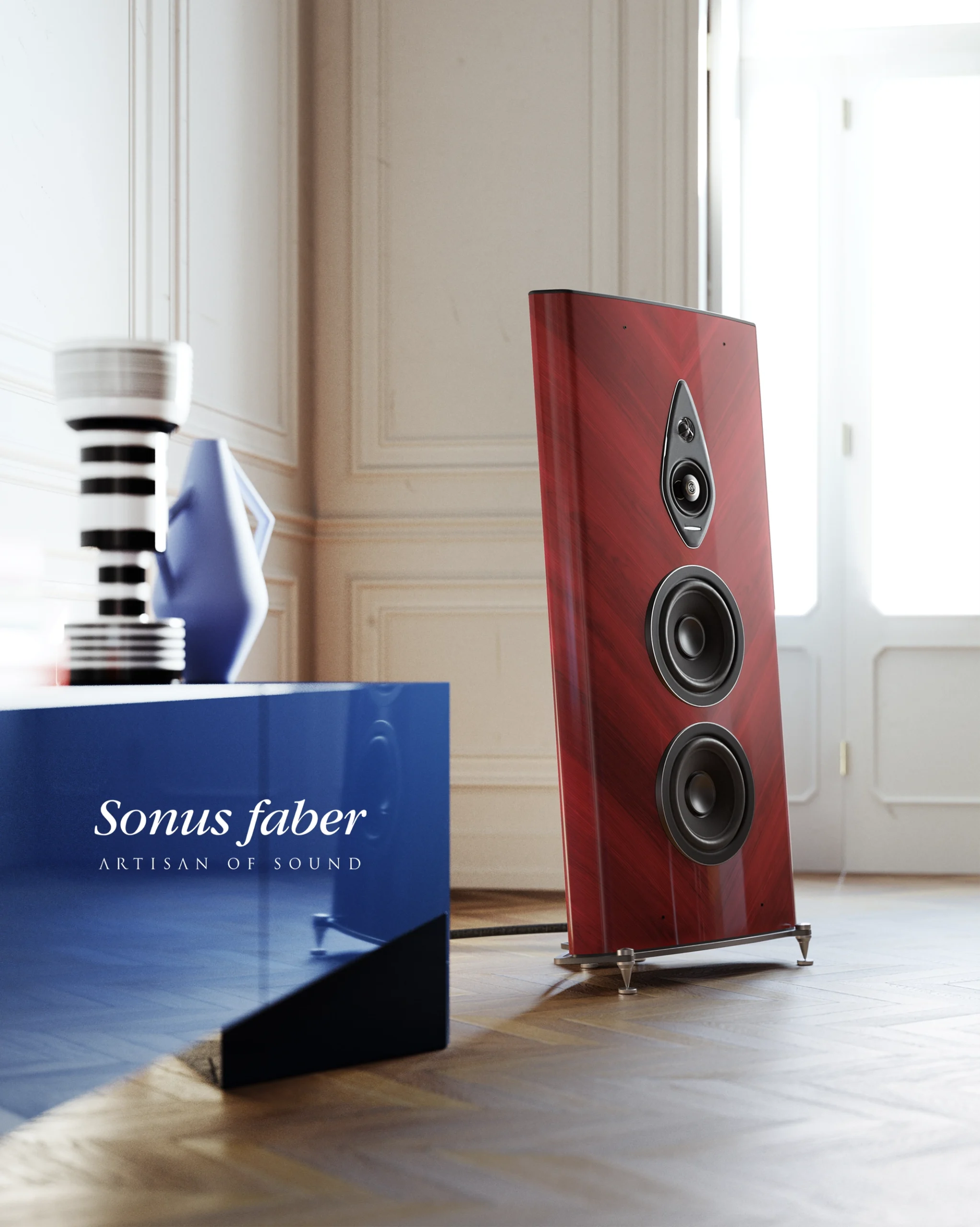 Red-lacquered Sonus Faber floorstanding speaker beside a blue plinth in a bright room with a parquet floor.