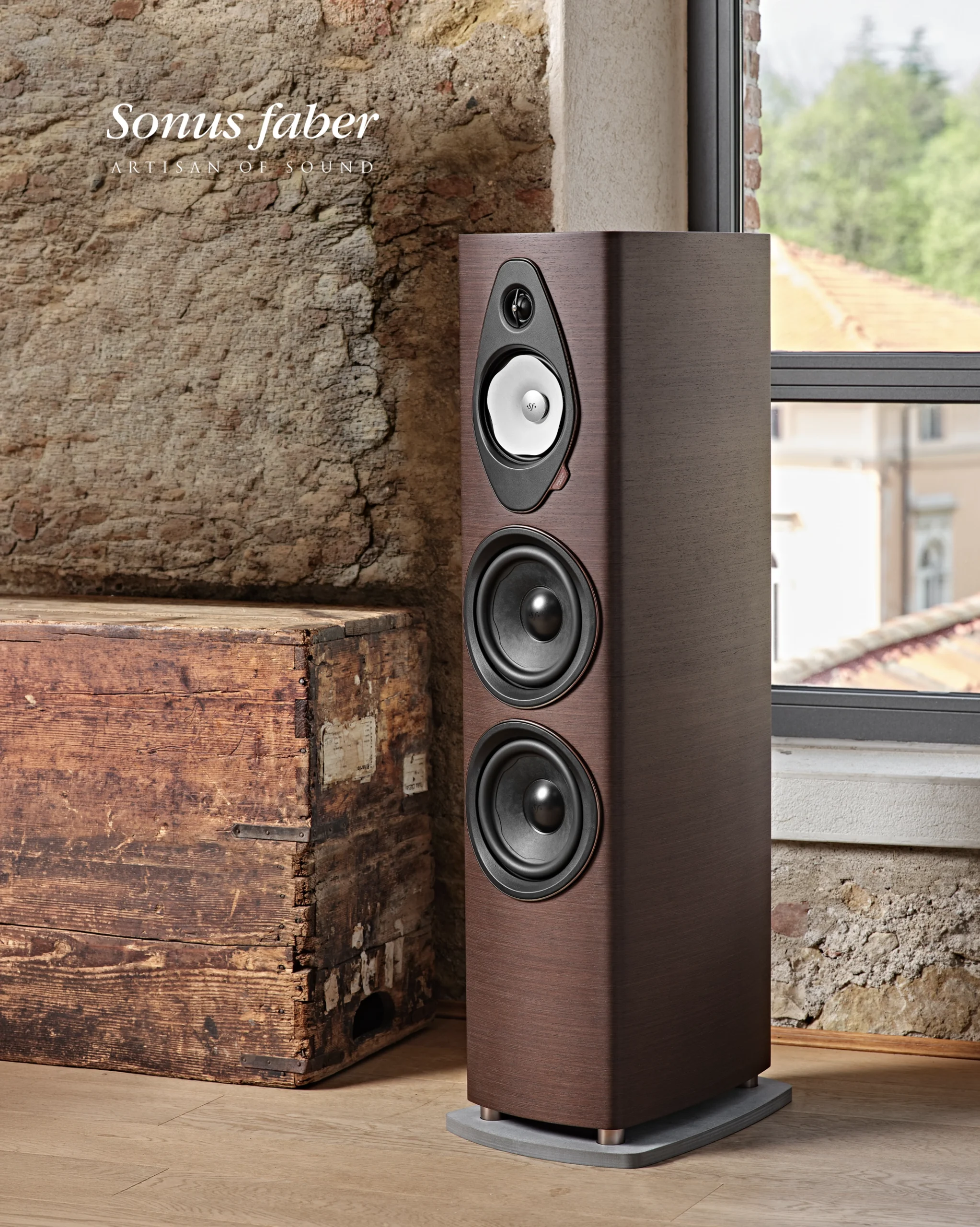 Tall Sonus Faber floorstanding speaker in dark wood against a stone wall and a window.