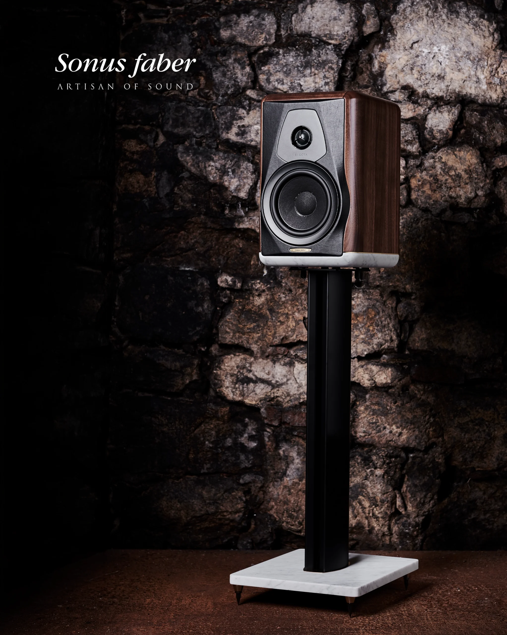 Sonus Faber bookshelf speaker in walnut on a black stand with a white stone base, against a dark stone wall.