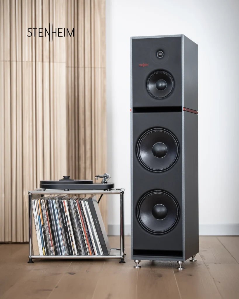 Tall Stenheim speaker with a black baffle and large bass drivers, beside a turntable and record rack in front of wooden slats.