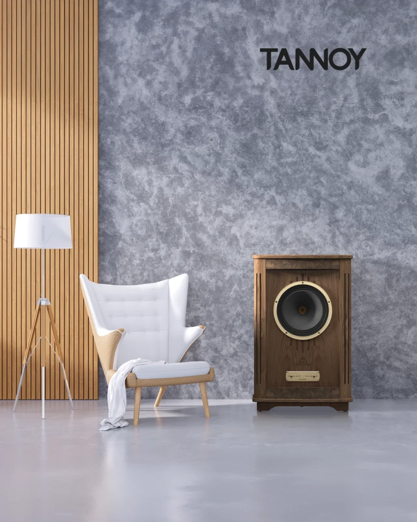 Large Tannoy floorstanding speaker in walnut with a gold driver ring, beside a white armchair and a grey textured wall.