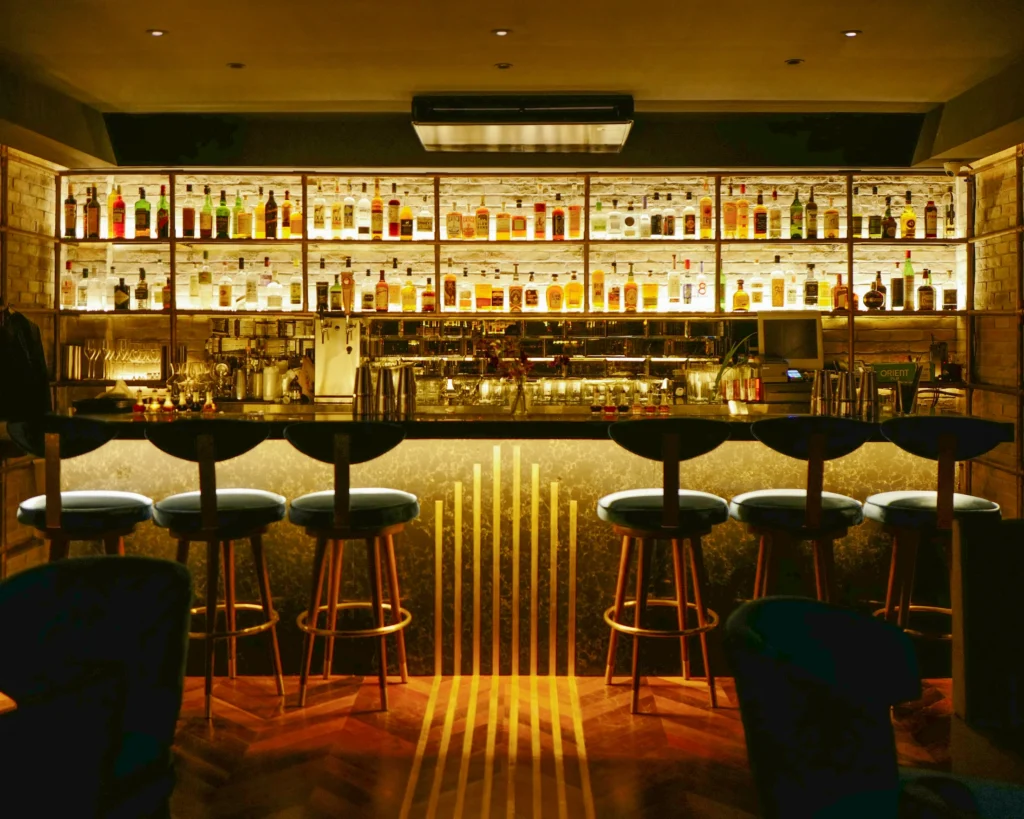 A softly lit cocktail bar with shelves of bottles, brass-trimmed bar stools and a herringbone timber floor
