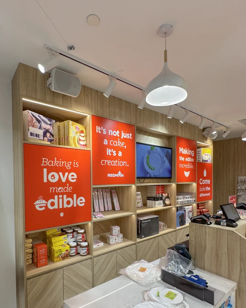 Retail shelving with a wall-mounted speaker on a track-lit wall, a small display screen and orange signage