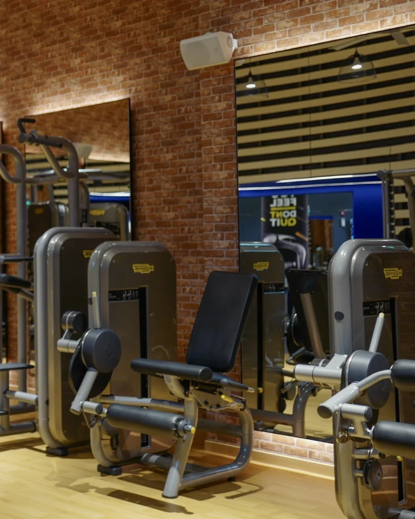A gym floor with strength machines against a brick wall, with a white speaker mounted above the mirrors