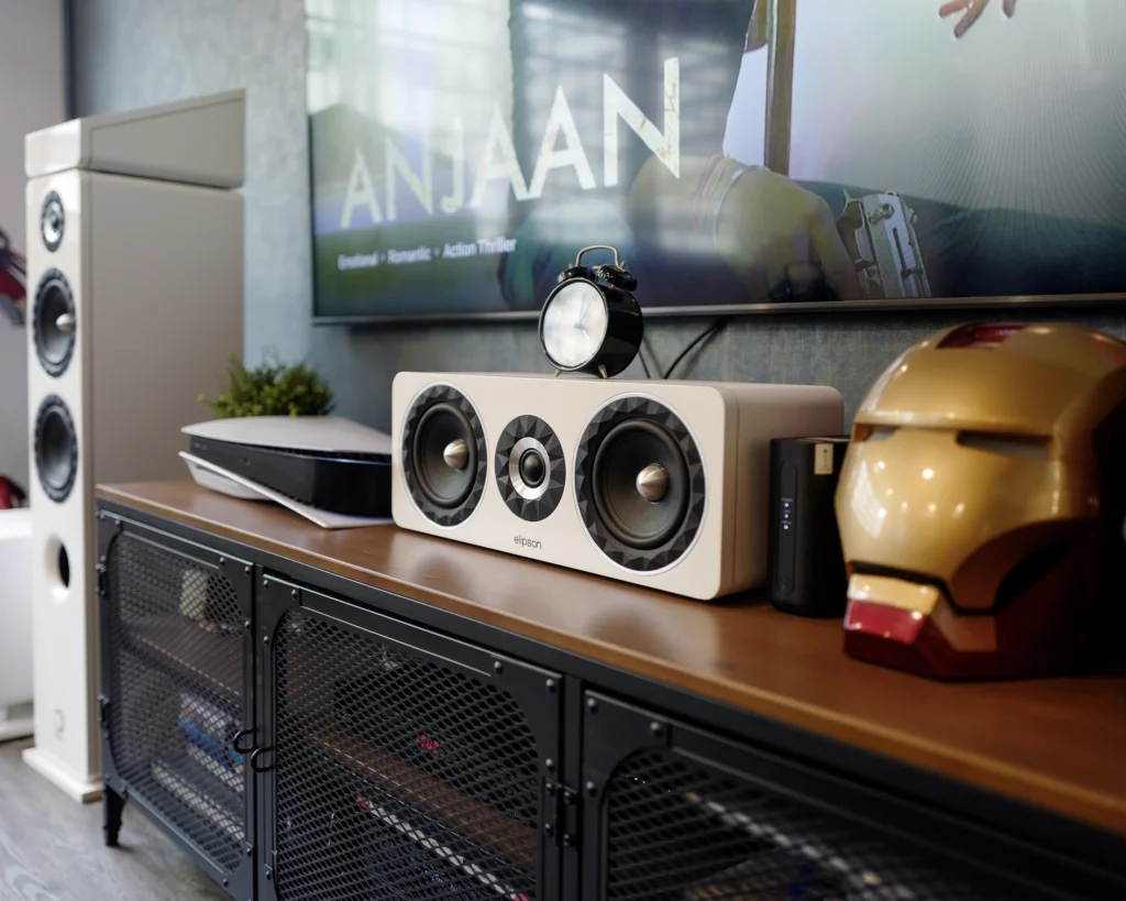 The white Elipson Prestige Facet centre speaker on a wooden media console, below a wall-mounted television.