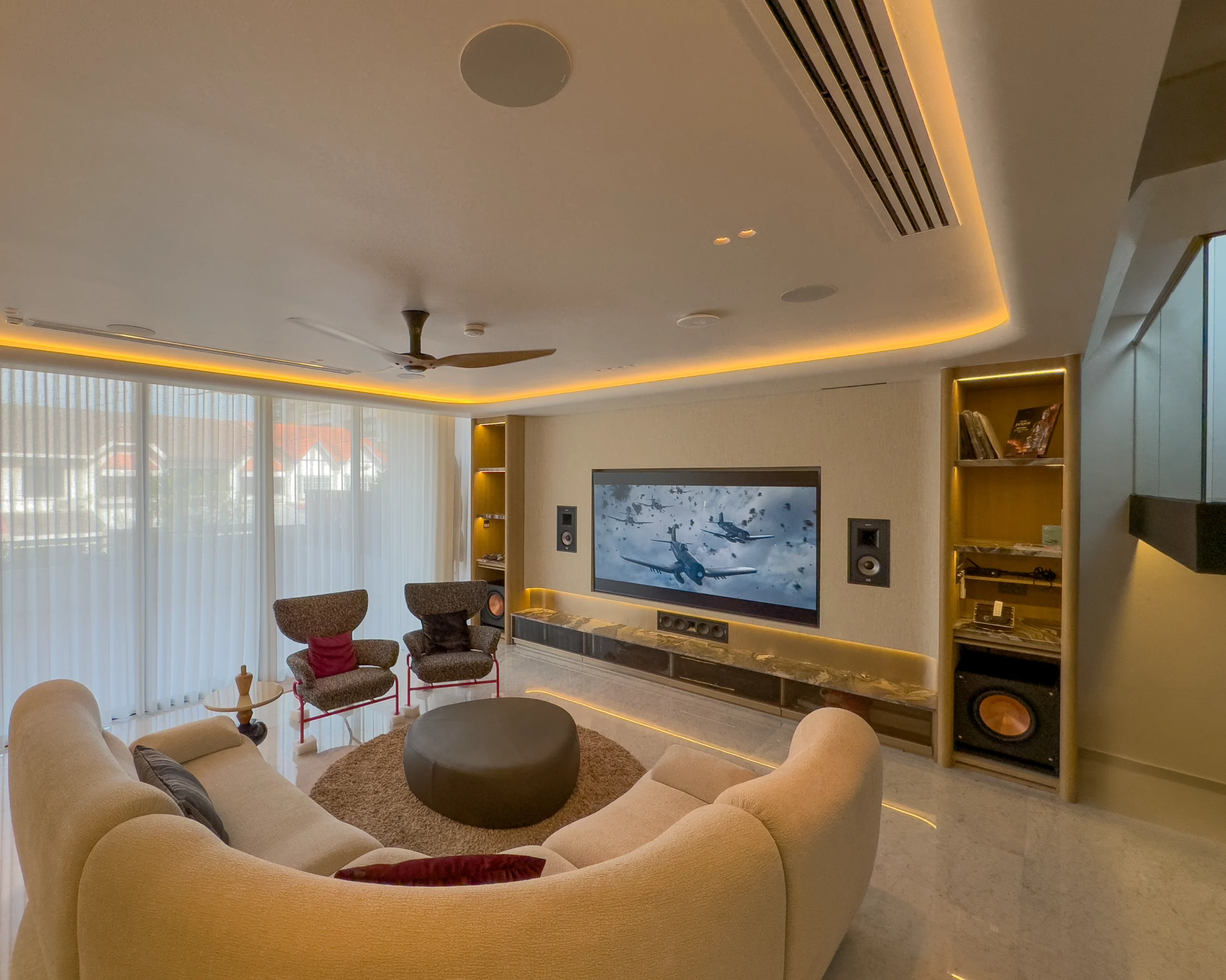 A living-room home theatre with a wide screen, in-wall speakers, a centre speaker, a subwoofer in a shelf and a curved sofa