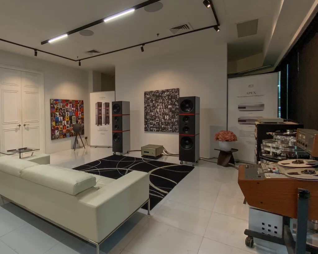 Listening room with two tall black floorstanding speakers, a cream sofa, framed record-sleeve artwork and a reel-to-reel tape machine