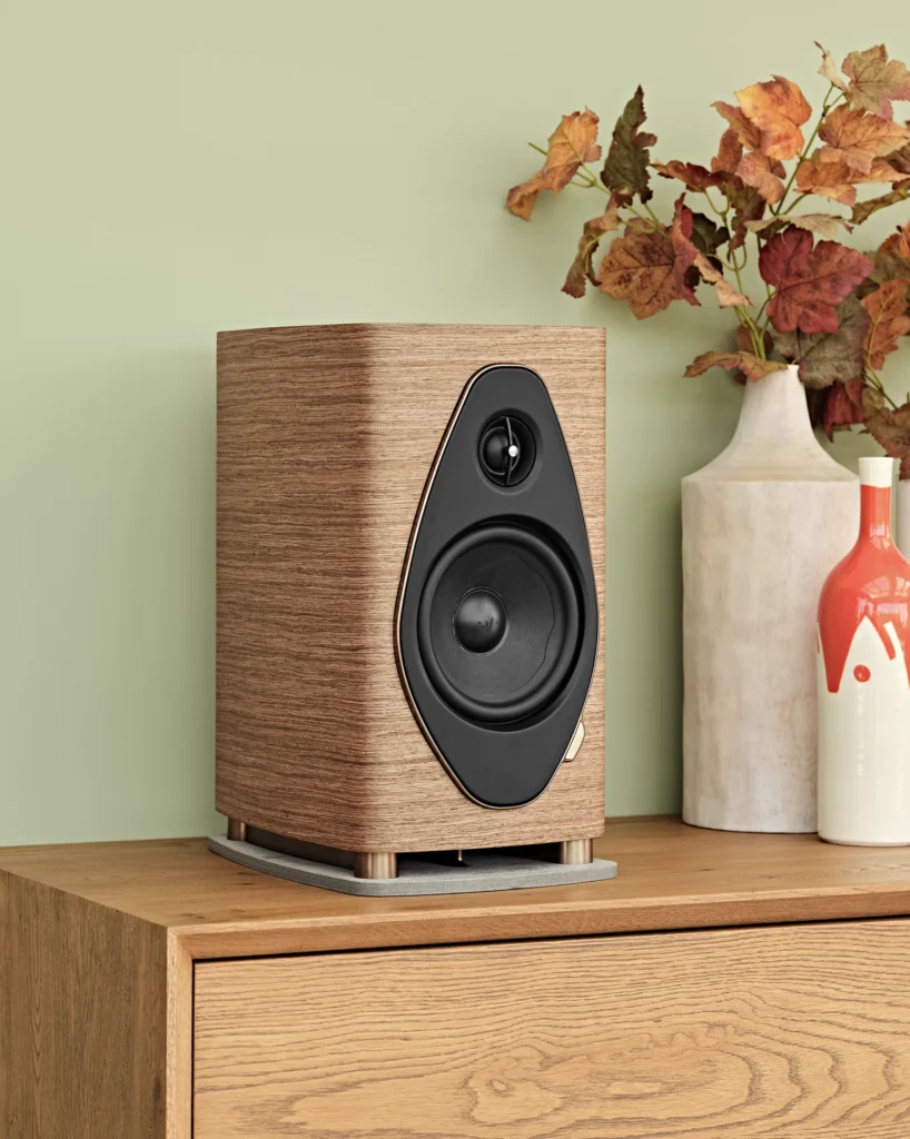A walnut Sonus Faber stand-mount loudspeaker on an oak sideboard against a sage green wall, beside pale ceramic vases.