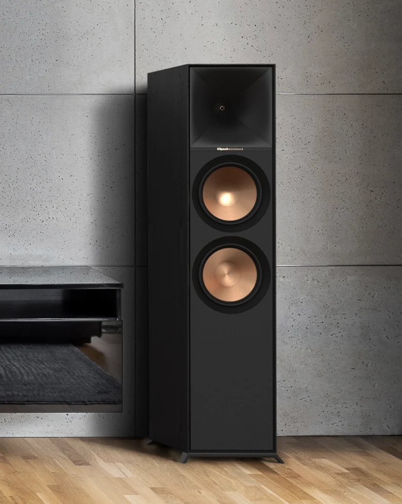 A black Klipsch Reference floorstanding loudspeaker with a horn tweeter and two copper woofers, against a concrete wall on a wood floor.