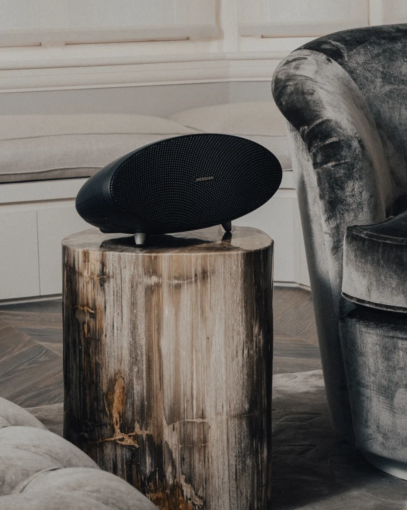 A black oval Meridian active loudspeaker on a petrified wood side table, next to a grey velvet armchair.