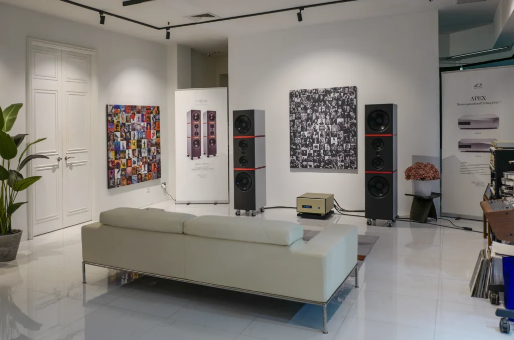 A white listening room with a cream sofa, two tall black floorstanding speakers and framed record-sleeve artwork