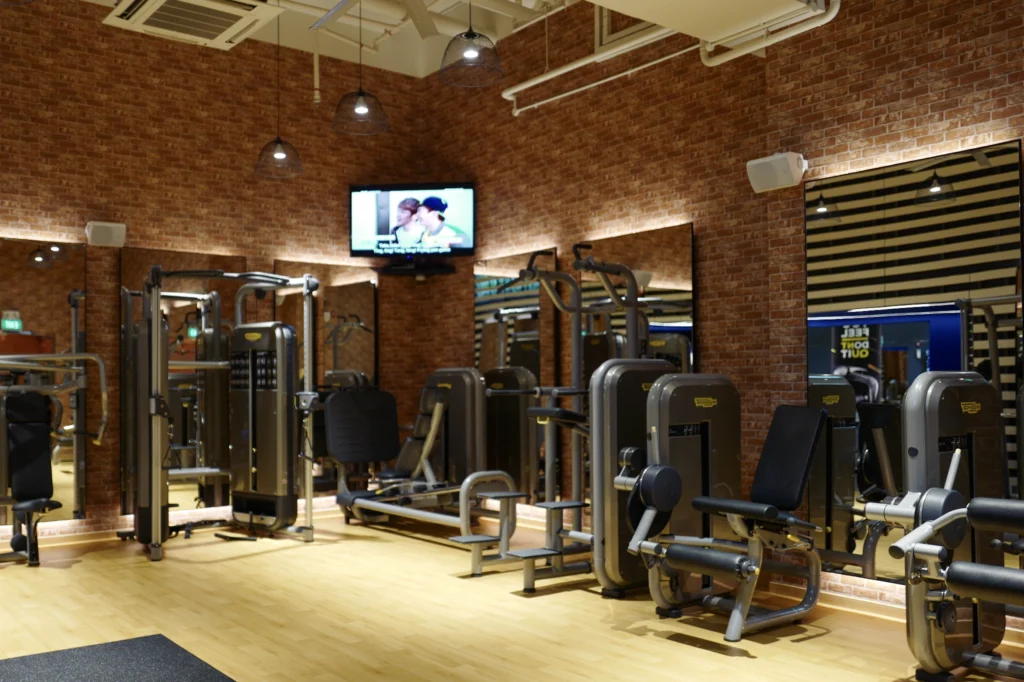 Condominium gym floor with weight machines, a brick wall, a wall-mounted TV and small speakers fixed high on the wall