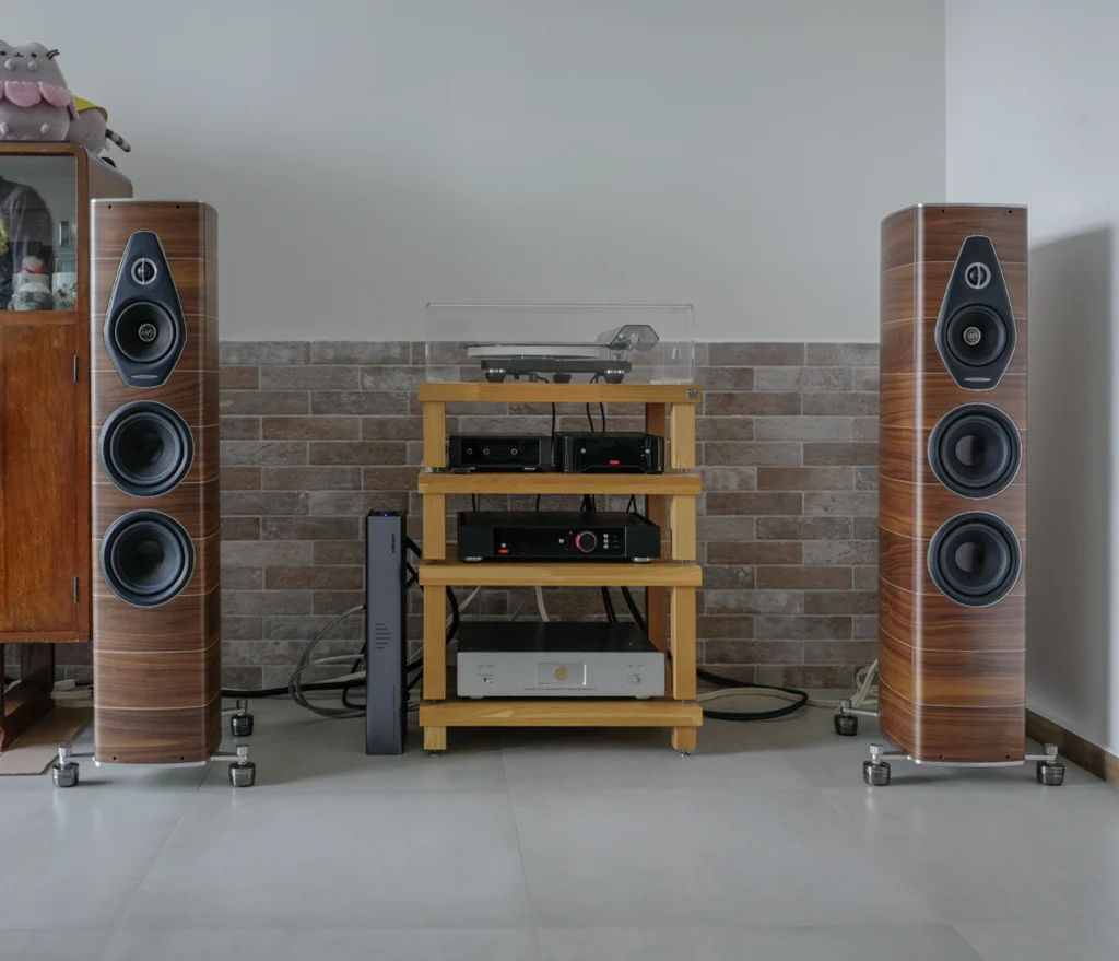 A pair of Sonus Faber floorstanding loudspeakers either side of a wooden rack holding a turntable and separates, against a brick wall.