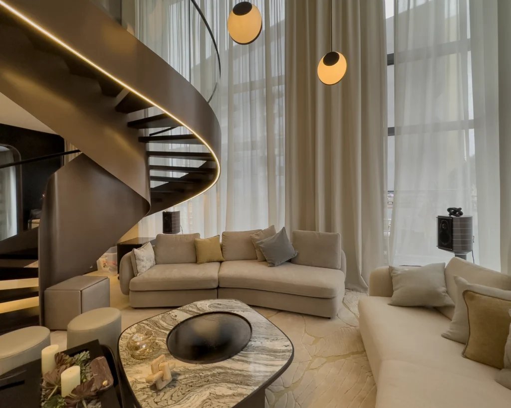Living room seating beneath a spiral staircase and full-height curtains, with a speaker in the far corner