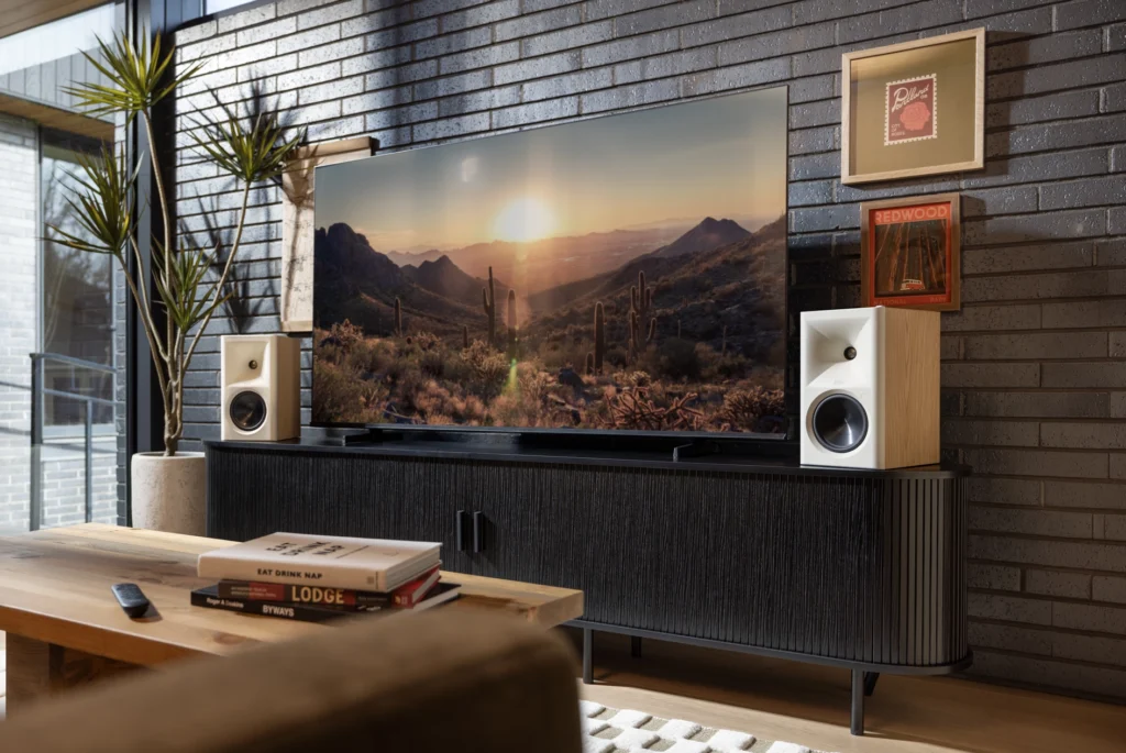 Two compact speakers in oak and white either side of a wall-mounted television above a black media cabinet in a living room