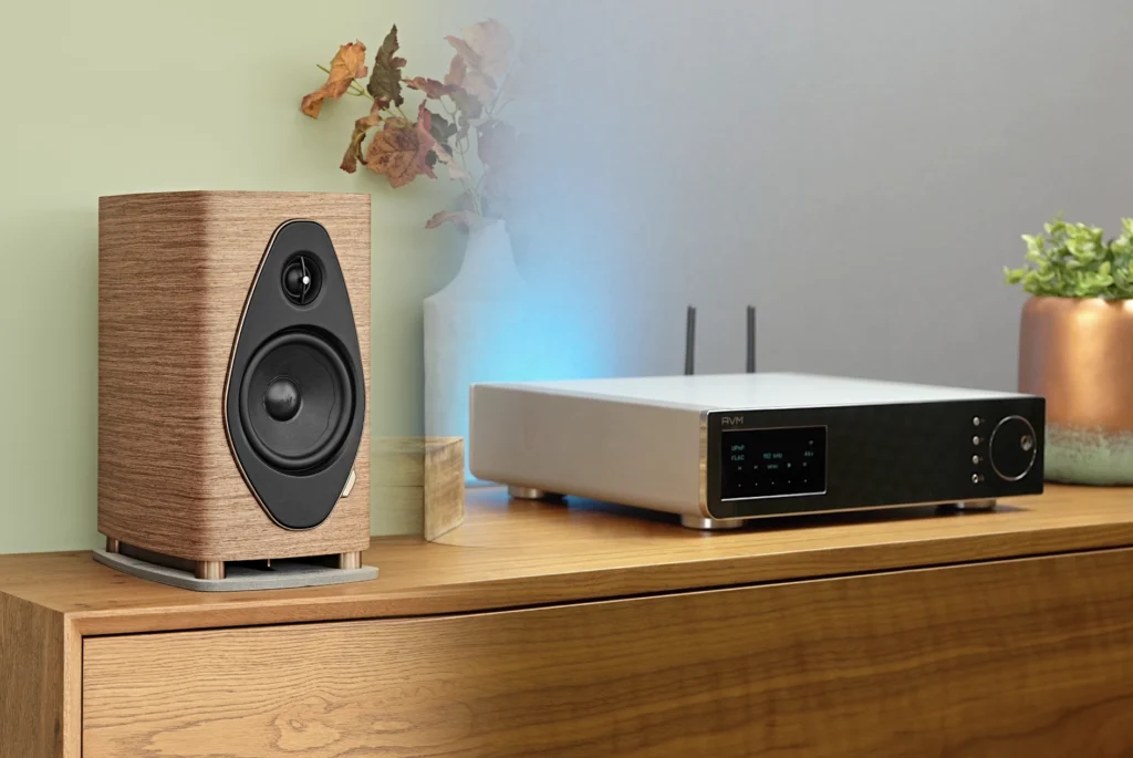 A walnut-finish bookshelf speaker beside a silver network streamer on a wooden sideboard