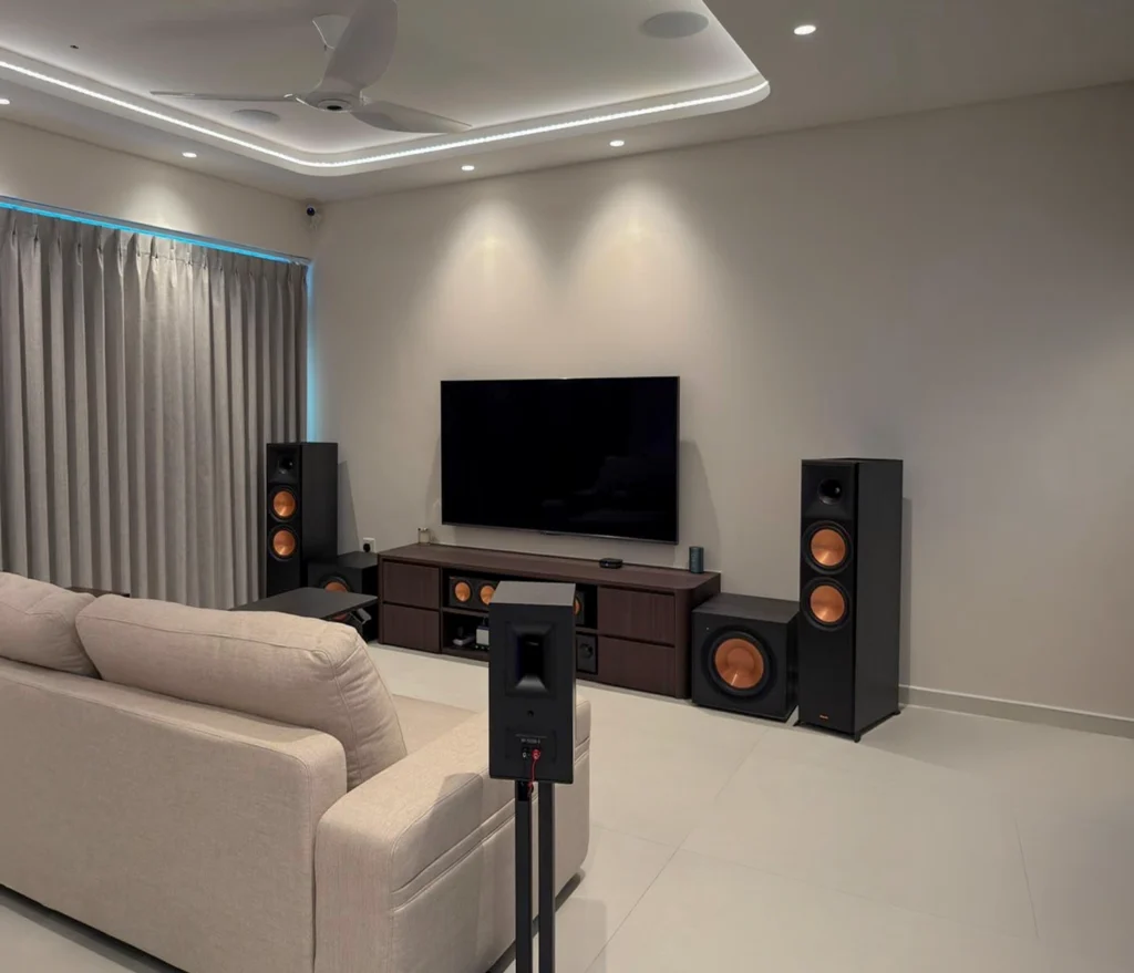 Condo living room from behind the sofa, with a surround on its stand, the speaker-lined front wall and a ceiling speaker in the cove.