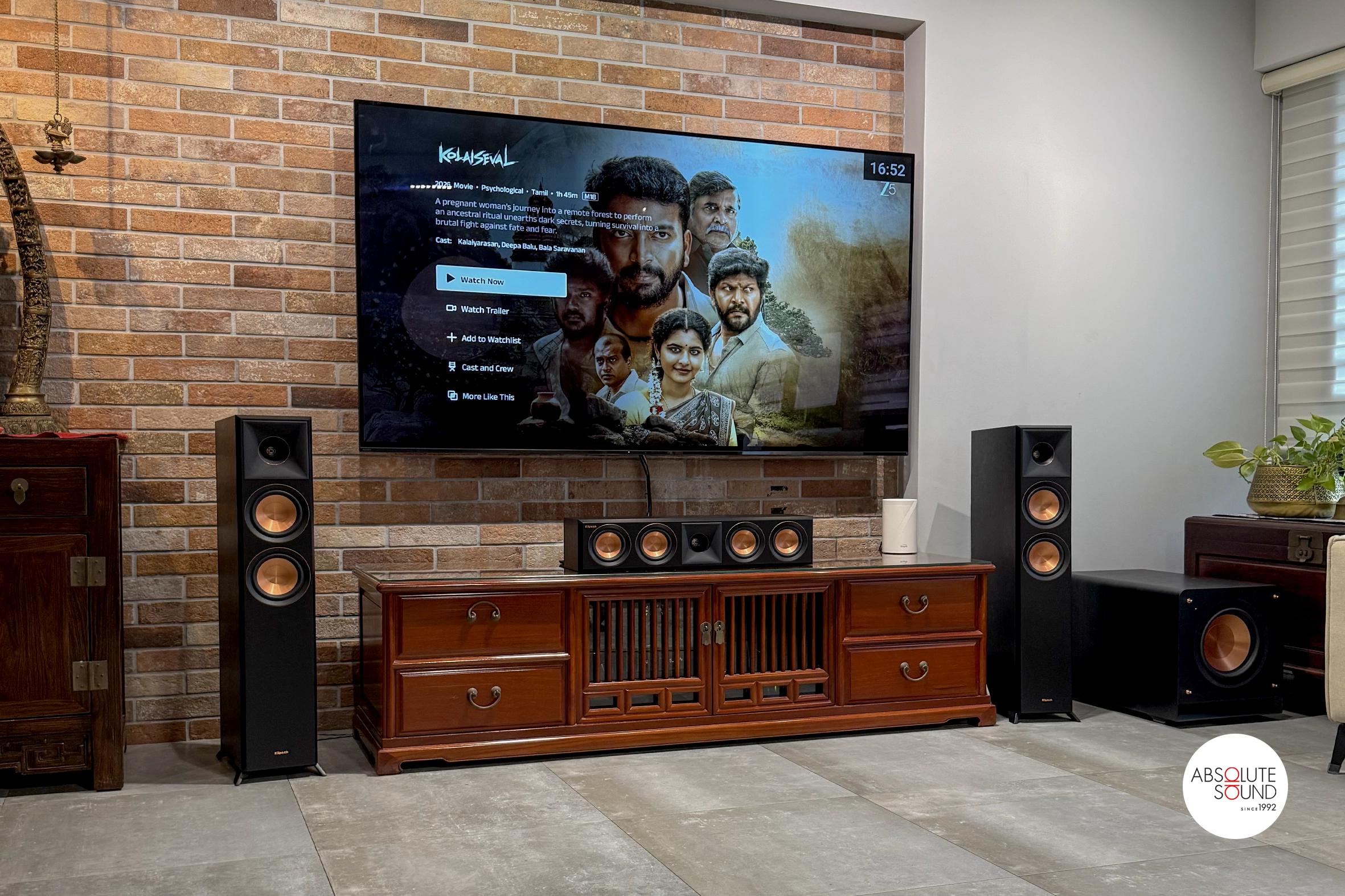 Front view of the ebony Klipsch centre speaker on a wooden cabinet below a wall-mounted TV, with a floorstander and subwoofer