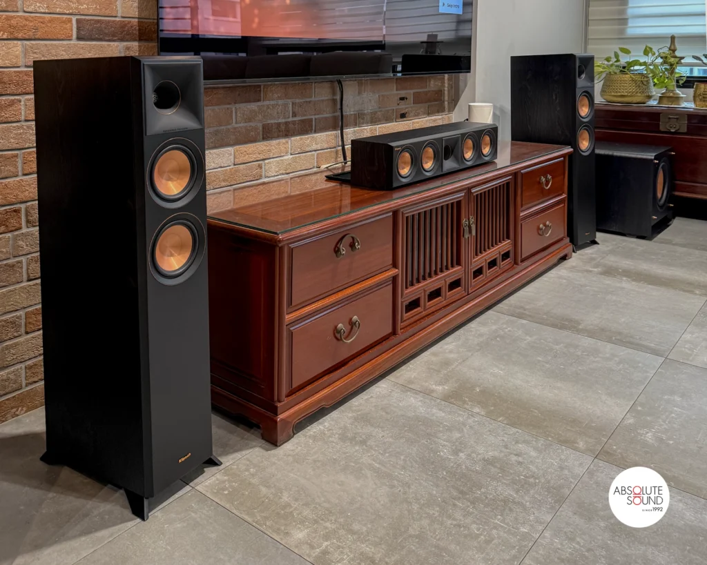 A black Klipsch floorstander in the foreground, the centre speaker on its cabinet, and the second floorstander and subwoofer.